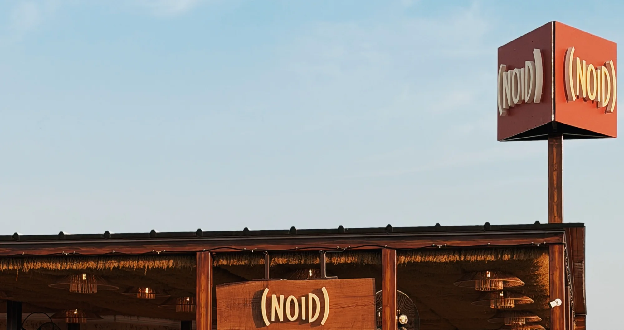 Павильон Coffee Noid в Sea Breeze: кубическая вывеска (NOID) из ржавой стали на мачте, крыша с камышовым свесом и деревянная табличка над входом
